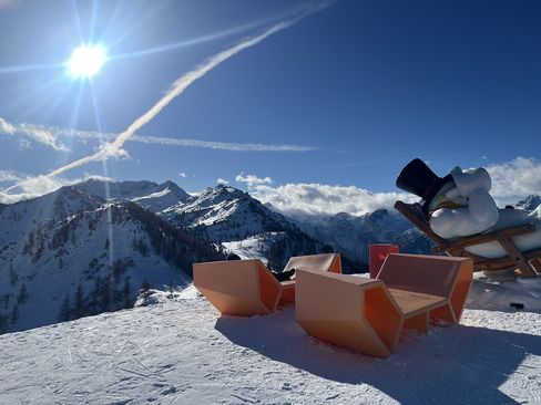 Enzi Sitzmöbel im Schnee auf einem alpinen Berggipfel, mit Blick auf verschneite Berge, strahlende Sonne und einen Schneemann im Hintergrund