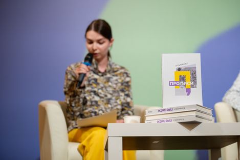 Woman holding a microphone and a notepad sitting on a chair behind a table with a stack of books, one standing upright with a visible title in Cyrillic script.