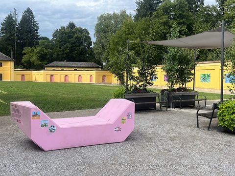 Pink Enzi in a park in front of a historic yellow building, surrounded by trees, plants and seating areas.