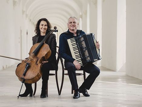 Zwei Personen sitzen Rücken an Rücken in einem hellen Gang, eine hält ein Cello, die andere ein Akkordeon.