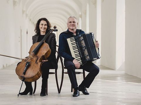 Two people sit back to back in a bright hallway, one holding a cello, the other an accordion.