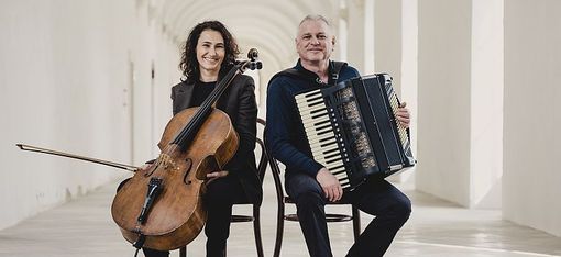 Zwei Personen sitzen Rücken an Rücken in einem hellen Gang, eine hält ein Cello, die andere ein Akkordeon.