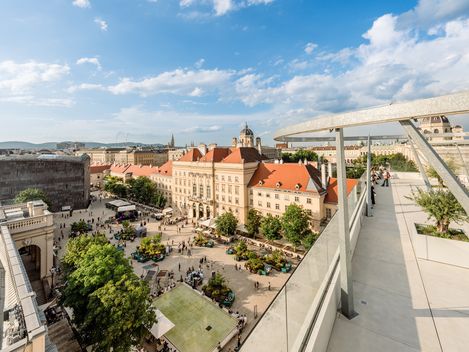 Blick von der Dachterrasse der MQ Libelle auf den belebten Haupthof des MuseumsQuartiers mit historischen Gebäuden, Bäumen und Menschen unter blauem Himmel mit Wolken.