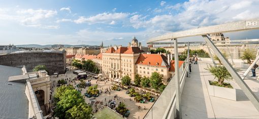 Blick von der Dachterrasse der MQ Libelle auf den belebten Haupthof des MuseumsQuartiers mit historischen Gebäuden, Bäumen und Menschen unter blauem Himmel mit Wolken.