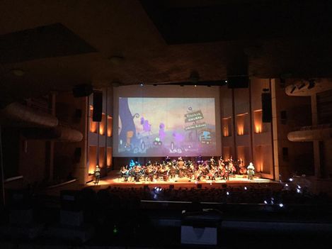 Orchestra on stage in front of a screen showing an animated projection in a dark concert hall.