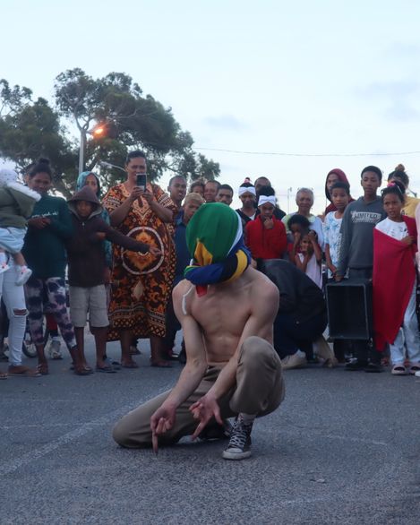 A shirtless muscular man wearing a colorful head covering kneels on the ground pointing downward with his fingers while a diverse group of people of various ages stands around him on a street.