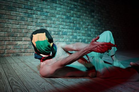 Person lying on their back on a wooden floor in front of a brick wall wearing a colorful head covering and pointing forward with an outstretched arm.