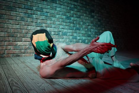 Person lying on their back on a wooden floor in front of a brick wall wearing a colorful head covering and pointing forward with an outstretched arm.