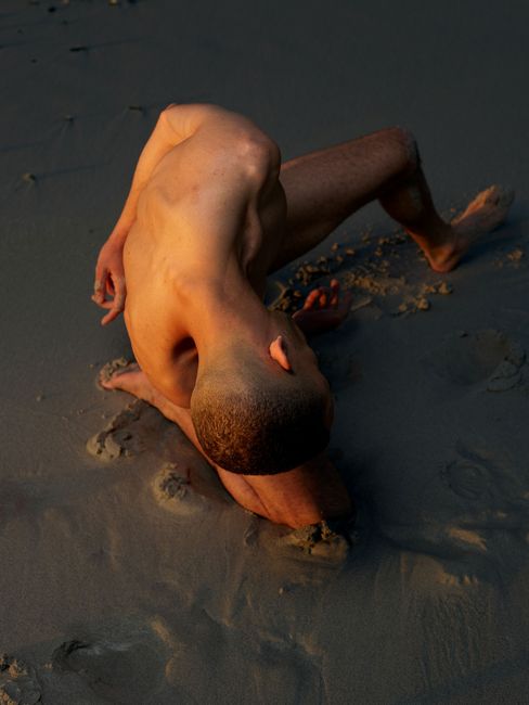 Top-down view of a shirtless person bending forward on sandy ground with head resting on forearm.