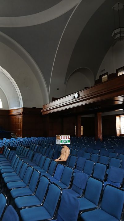 A naked man sits alone in a large hall with rows of blue chairs and high vaulted ceilings.