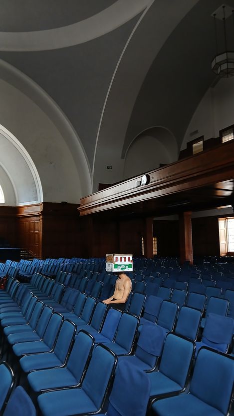 A naked man sits alone in a large hall with rows of blue chairs and high vaulted ceilings.
