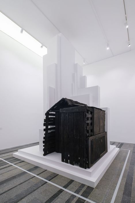 Sculpture of a small house made of dark wooden slats in a white exhibition room.