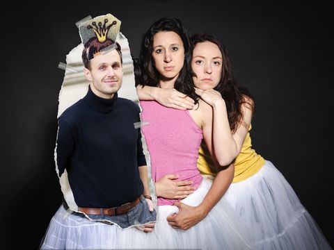 Das Bild zeigt drei Personen, die dicht beieinander stehen, mit zwei Frauen und einem Bild eines Mannes vor der dritten Person. Das Bild scheint aus einem Papier herausgerissen worden zu sein. Auf seinem Kopf ist eine Krone mit Klebeband befestigt.