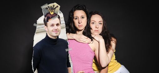 Das Bild zeigt drei Personen, die dicht beieinander stehen, mit zwei Frauen und einem Bild eines Mannes vor der dritten Person. Das Bild scheint aus einem Papier herausgerissen worden zu sein. Auf seinem Kopf ist eine Krone mit Klebeband befestigt.