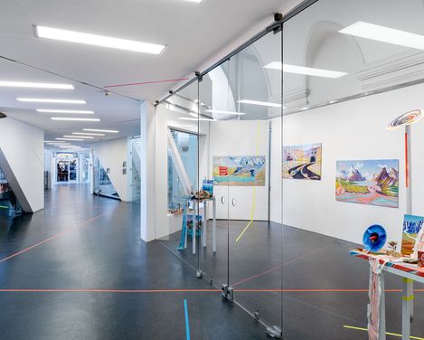 Blick in einen modernen Ausstellungsraum mit Glaswänden, auf denen bunte Gemälde und Kunstwerke zu sehen sind, sowie roten und blauen Linien auf dem Boden und an den Wänden.