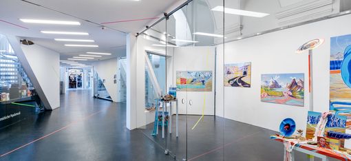 Blick in einen modernen Ausstellungsraum mit Glaswänden, auf denen bunte Gemälde und Kunstwerke zu sehen sind, sowie roten und blauen Linien auf dem Boden und an den Wänden.