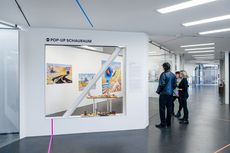 Drei Personen betrachten in einem modernen Ausstellungsraum mit weißer Wand und großen Fenstern bunte Gemälde an der Wand