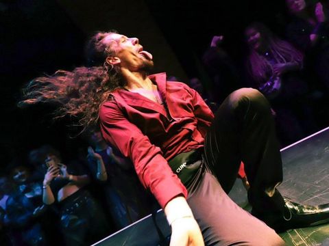 Man with long hair and red shirt sitting on a stage floor sticking out his tongue