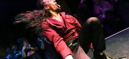 Man with long hair and red shirt sitting on a stage floor sticking out his tongue