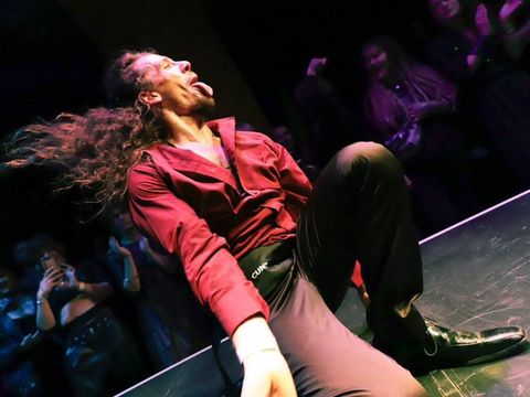 Man with long hair and red shirt sitting on a stage floor sticking out his tongue