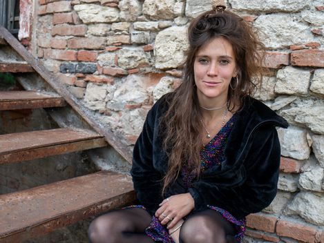 Woman with long curly hair sitting on a stair step in front of a stone wall wearing a black jacket and patterned dress