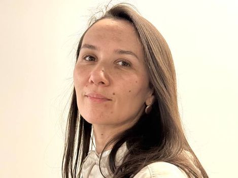 Frau mit langen braunen Haaren trägt eine helle Bluse und steht vor hellem Hintergrund.