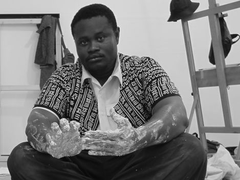 Person sitting cross-legged on the floor with paint-covered hands wearing a patterned shirt over a white shirt.