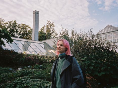 Person with pink hair wearing a green sweater and dark coat standing in front of a greenhouse and trees.