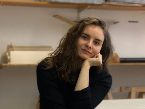 Person mit langen Haaren sitzt mit verschränkten Beinen und stützt den Kopf mit der Hand auf einem Stuhl in einem Raum mit Regalen und Tischen