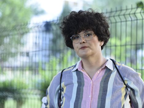 Porträt einer Frau mit lockigem Haar und Brille vor einem unscharfen Zaun im Freien.