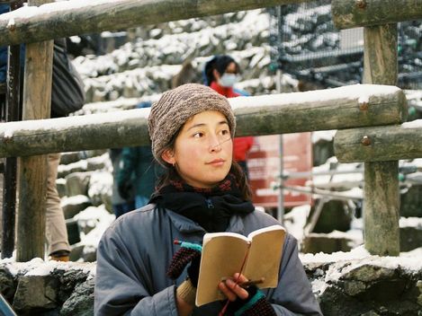 Person wearing a knit hat and gloves holding a notebook in front of a snow-covered wooden railing and stone wall.
