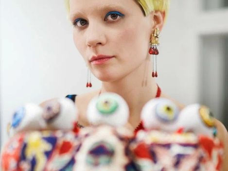 Porträt einer Person mit kurzem blondem Haar, blauem Augen-Make-up und auffälligem Schmuck.