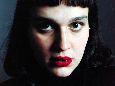 Portrait of a person with dark hair, straight short bangs, and red lipstick against a dark background