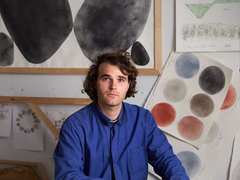 Junger Designer im Atelier, sitzt an einem Tisch mit gelb-weißen Muster-prints, im Hintegrund eine Pinnwand voller Skizzen.