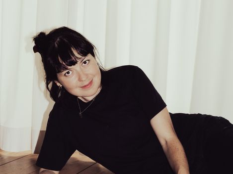 Person with tied-back hair wearing a black t-shirt lying on the floor petting a light-colored cat in front of a white curtain.