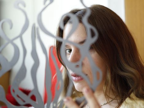 Person with shoulder-length hair wearing a yellow shirt in front of a glass panel with an abstract pattern.