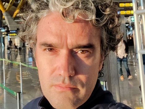 Portrait of a man with curly gray hair and dark jacket in a bright modern hall with yellow steel beams.