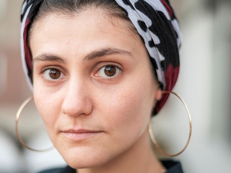 Person mit gemustertem Kopftuch und großen Creolen, trägt ein dunkles Hemd mit offenem Kragen.