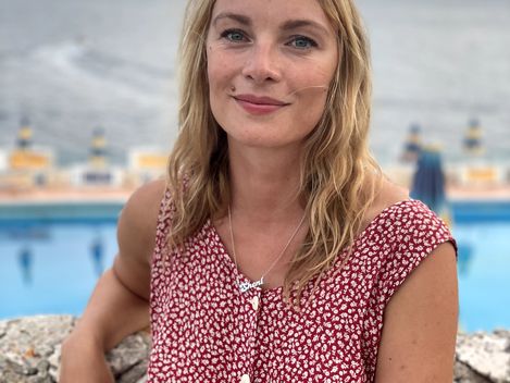 Woman with shoulder-length hair wearing a red patterned dress leaning on a stone wall with a pool and sea in the background.