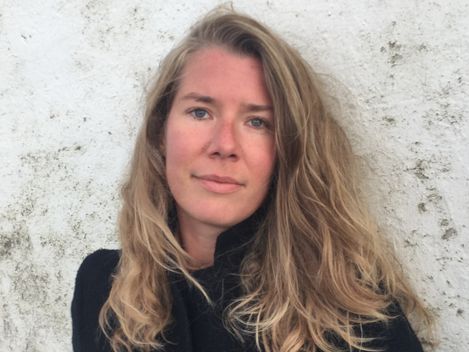 Person mit langen, lockigen Haaren vor einer hellen, strukturierten Wand.