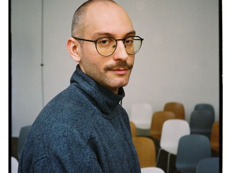 Porträt einer Person mit Brille und grauem Pullover vor einer Wand und mehreren Stühlen in verschiedenen Farben.