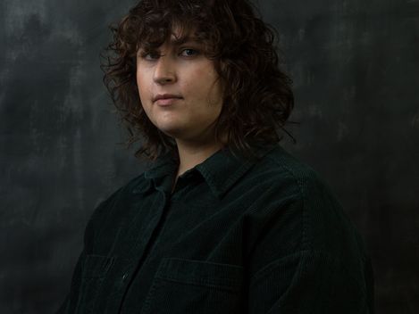 Person with curly hair wearing a shirt against a dark background.