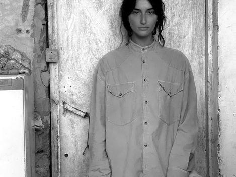 Person with long hair and cap standing in front of an old metal door in a dilapidated room.