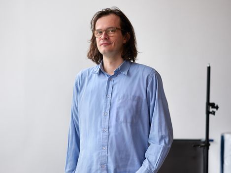 Standing person wearing a blue shirt and black pants in front of a white wall and a table with small objects.