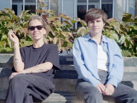 Two people sitting on a park bench in front of a building with large windows and bushes in the background.