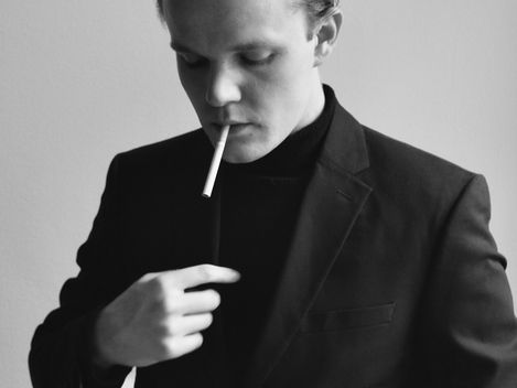 Black-and-white portrait of a man in a suit jacket holding the jacket with one hand and pointing to his chest with the other.