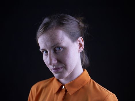Person with tied-back hair wearing an orange blouse against a black background.