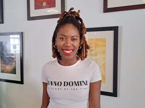 Person wearing a white T-shirt with the text "ANNO DOMINI SIGNS OF THE TIMES" standing in front of a wall with framed certificates.