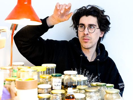 Person wearing a dark hoodie sits behind a table with many small jars and raises their right hand beside their head.