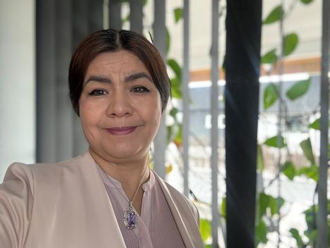 Woman with tied-back hair wearing a light blouse and a necklace with a pendant standing in front of a window with vertical blinds and green plants.